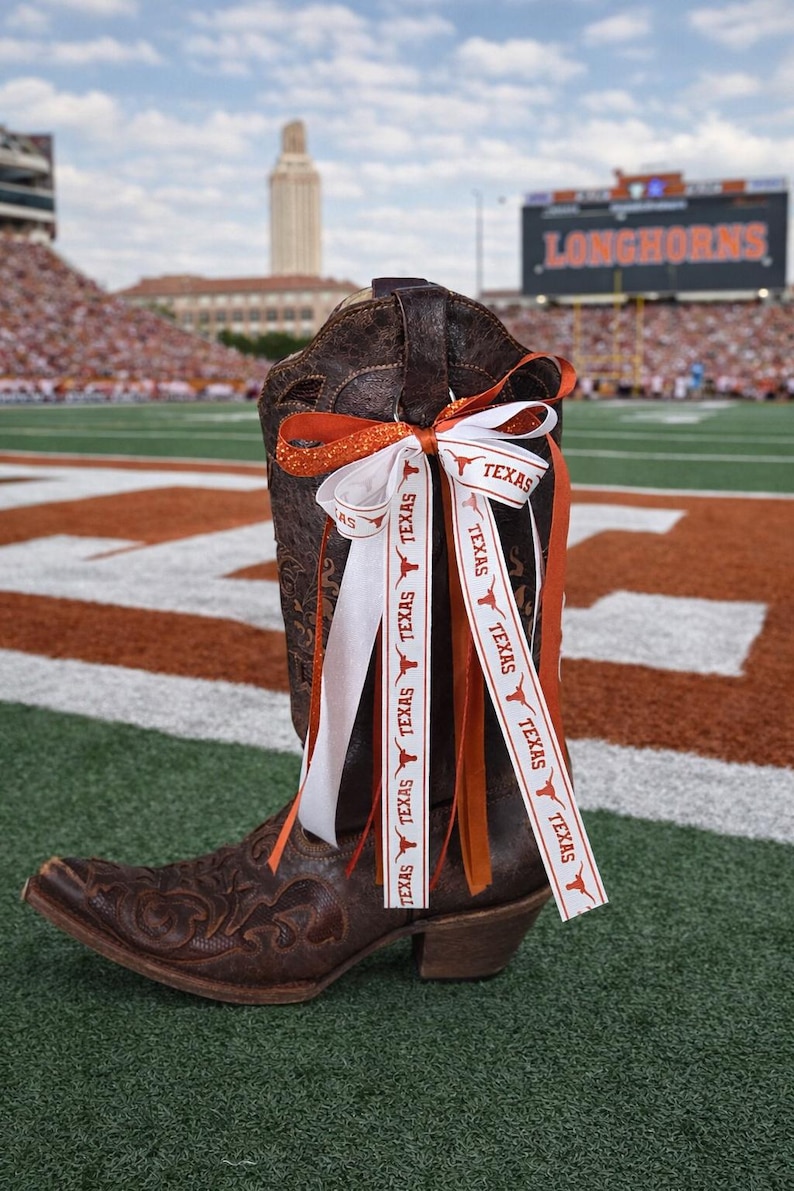 May include: A brown leather cowboy boot with decorative stitching, adorned with orange and white ribbons. The ribbons are printed with the word "TEXAS" and a Longhorn logo. The boot is on a football field.