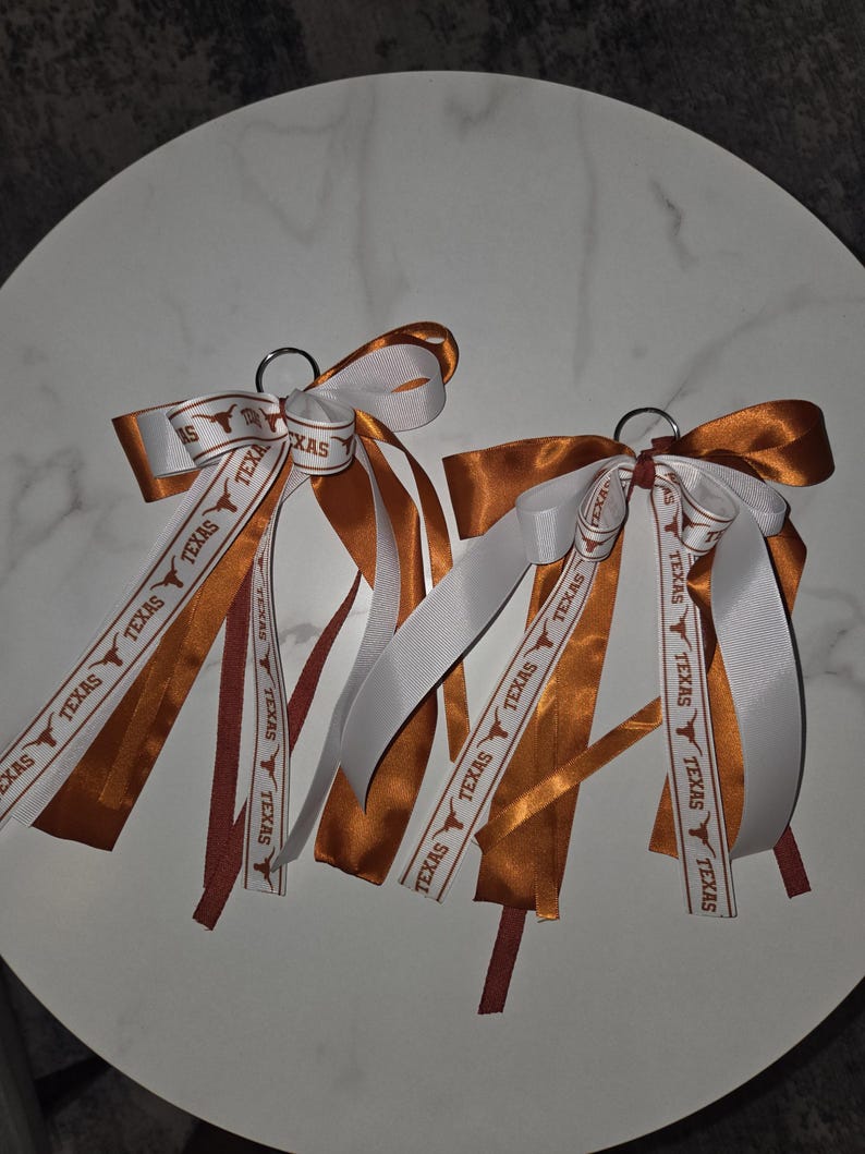 May include: Two keychains with orange, white, and maroon ribbons. The ribbons are printed with the word "TEXAS" and a Longhorn logo. The keychains are likely used for school spirit or as a souvenir.