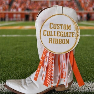May include: A white cowboy boot with a round sign that reads "CUSTOM COLLEGIATE RIBBON." The sign is decorated with gold glitter and orange and white ribbons. The ribbons are printed with the word "Tennessee."