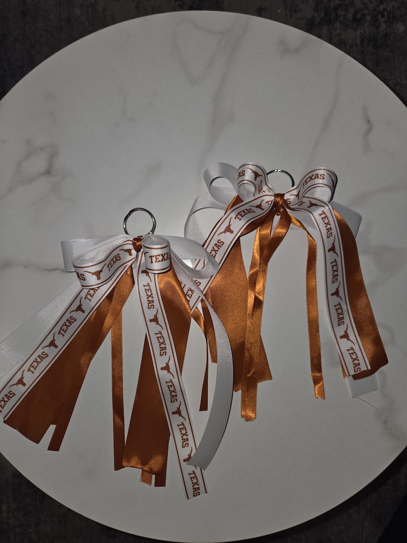 May include: Two keychains with white and burnt orange ribbons. The ribbons are printed with the word "TEXAS" and a Longhorn logo. The keychains are on a white marble surface.
