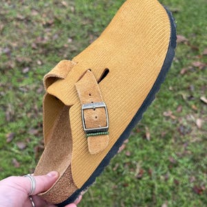 May include: Mustard yellow corduroy clog with a black sole and a buckle closure. The shoe has a cork footbed and a strap with a silver buckle. The shoe is designed for comfort and casual wear.