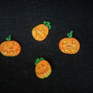 May include: Four orange pumpkin decorations with glitter and green stems are arranged on a black surface. Each pumpkin has a carved face with triangular eyes and a smiling mouth, perfect for Halloween.