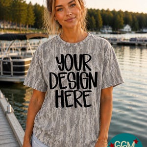 May include: A short-sleeved t-shirt with a grey and white camouflage pattern. The front of the shirt features the black text "YOUR DESIGN HERE" in a bold, rounded font. The garment has a crew neckline and a relaxed fit, constructed from fabric.