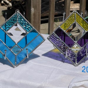 May include: Two stained glass diamond-shaped suncatchers. One is blue and turquoise, the other is purple, yellow, and black. Each has a silver metal frame and a small hook for hanging. The numbers 19 and 20 are written on the surface.