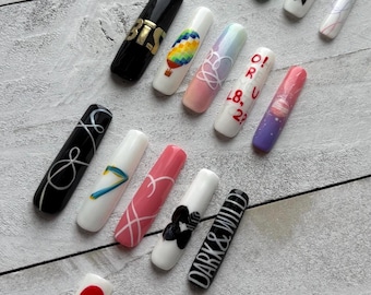 May include: A collection of sixteen long, rectangular press-on nails featuring various designs including the text LUV AFFAIR, BE, BTS, O!RUL8,2?, and DARK&WILD. The nails display diverse patterns such as a hot air balloon, a planet, a number seven, and abstract line art in colors including black, white, pink, green, and blue.