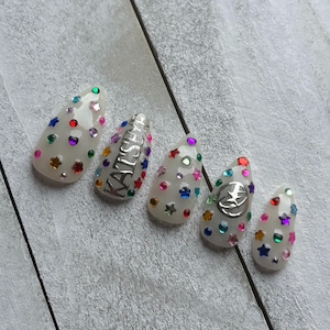 May include: Five off-white almond-shaped press-on nails decorated with colourful star and circular rhinestones. One nail features the silver text KATSEYE, while another displays a circular silver emblem with a star design in the centre.