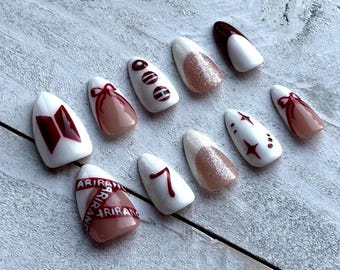 May include: A collection of ten artificial nails, each painted with a unique design. The nails feature a variety of colors, including white, brown, and glitter accents. Some nails have bow, star, and number designs, while others display text.