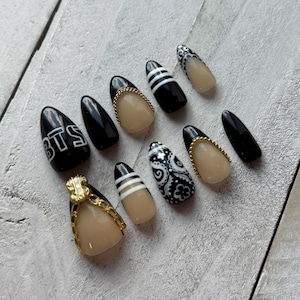 May include: A set of ten almond-shaped false nails. Designs include solid black, black with white stripes, a paisley pattern, and a nude base with gold chain and gem accents. One nail features the letters "BTS" in white.