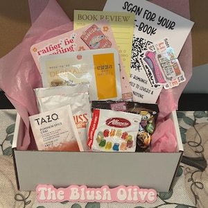 May include: A gift box filled with various items, including a Tazo Pumpkin Spice Chai tea bag, a face mask, gummy bears, and stickers. The box contains pink tissue paper and a sign that reads "The Blush Olive."
