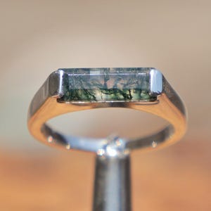 May include: A silver-toned metal ring featuring a rectangular, horizontally set moss agate gemstone. The stone is translucent with dark green, branch-like inclusions visible throughout its surface, held securely by a smooth, polished metal band.