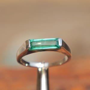 May include: A silver-toned metal ring featuring a single, horizontally set, rectangular baguette-cut green gemstone. The stone is securely held within a smooth, polished metal band that tapers slightly toward the base of the setting.