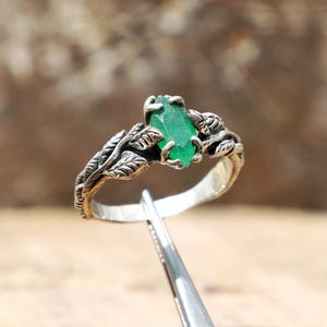 Puede incluir: Un anillo de plata con una piedra preciosa de esmeralda verde. El anillo presenta un diseño de hojas alrededor de la piedra preciosa y en la banda. El anillo se muestra sobre una superficie de madera.