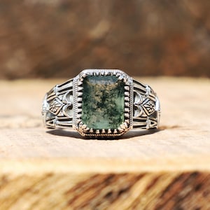 Puede incluir: Anillo de plata con una piedra verde musgo de forma rectangular. El anillo tiene una banda grabada con diseños florales y geométricos. La piedra está engastada en un marco rectangular con un borde de cuentas, creando un estilo vintage.