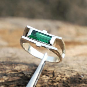 May include: A silver ring with a rectangular, emerald-green gemstone. The ring has a modern, geometric design with a band that splits into two parallel lines on either side of the gemstone. The ring is held by a pair of tweezers against a wooden background.
