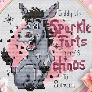 Puede incluir: Un bastidor de bordado de punto de cruz con un burro de dibujos animados y el texto "Giddy Up Sparkle Farts There's Chaos To Spread." El burro es gris y negro, sobre un fondo rosa. El bordado está en un bastidor de madera.