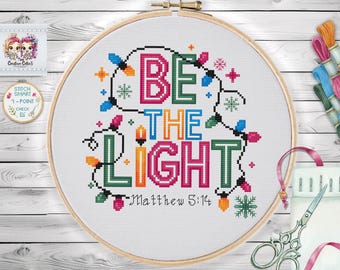 Może przedstawiać: Obręcz do haftu krzyżykowego z napisem "BE THE LIGHT" w kolorowych literach, otoczona łańcuchami świetlnymi i płatkami śniegu. Tekst "Matthew 5:14" znajduje się poniżej. Haft jest umieszczony w drewnianej obręczy.