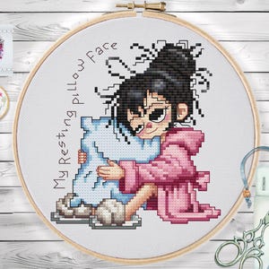 Op de afbeelding: Kruissteek borduurring met een cartoonmeisje in een roze badjas die een blauw kussen omhelst. De tekst "My Resting pillow Face" is rond de afbeelding gestikt. Houten achtergrond.