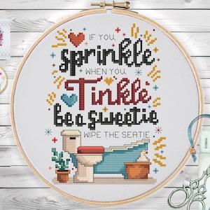 Op de afbeelding: Een kruissteek borduurring met de tekst "If you sprinkle when you tinkle be a sweetie wipe the seatie." Het ontwerp bevat een toilet, een bad en decoratieve elementen in zwart, rood, blauw en geel.