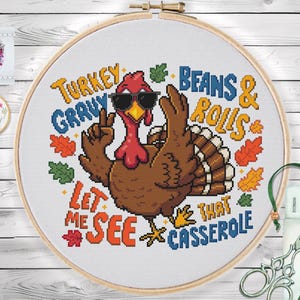 May include: A cross-stitch design in a wooden hoop features a cartoon turkey wearing sunglasses. The text reads "Turkey Gravy, Beans & Rolls, Let Me See That Casserole" in colorful letters. Autumn leaves surround the turkey.