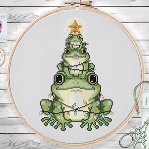 Könnte beinhalten: Kreuzstichmuster eines Weihnachtsbaums aus grünen Fröschen, verziert mit bunten Lichtern und einem goldenen Stern. Das Design befindet sich in einem hölzernen Stickrahmen, mit zusätzlichem Bastelzubehör.