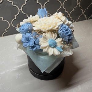 May include: A floral arrangement in a black round box. The bouquet features white and blue soap flowers, including daisies, peonies, and other blooms. The flowers are arranged with small, dried filler flowers and wrapped in light blue tissue paper.