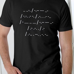 May include: Black t-shirt featuring white Morse code symbols. The text, composed of dots, dashes, and forward slashes, is printed across the chest. The t-shirt has a crew neck and is likely made of cotton.