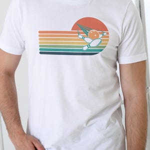 May include: White t-shirt with a retro bowling design. The graphic shows a bowling ball, pins, and a stylized sunset with horizontal stripes in orange, yellow, green, and blue. The design has a vintage aesthetic.