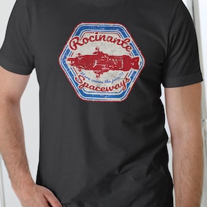 May include: A dark grey t-shirt with a distressed graphic. The design is a red and white hexagon with the words "Rocinante Speedway" and a red spaceship illustration. The text "Here comes the juice!!!" is also included.
