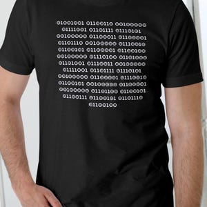 May include: Black t-shirt featuring white binary code text. The text reads: "01001001 01100110 00100000 01111001 01101111 01110101 00100000 01100011 01100001 01101110 00100000 01110010 01100101 01100001 01100100 00100000 01110100 01101000 01101001 01110011 00100000 01111001 01101111 01110101 00100000 01100001 01110010 01100101 00100000 01100001 00100000 01101100 01100101 01100111 01100101 01101110 01100100".