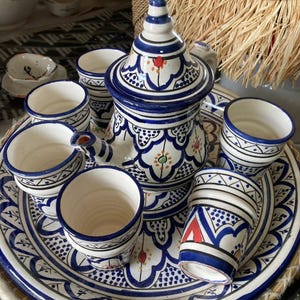 Peut inclure: Service à thé en céramique blanche et bleue, comprenant une théière, six petites tasses et un plateau de service. L'ensemble présente des motifs bleus détaillés avec des accents rouges et verts. Le service à thé est présenté sur un plateau tressé.