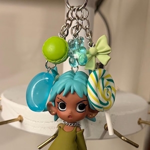 May include: A whimsical keychain featuring a doll with blue hair and a green outfit, adorned with charms like a macaron, a bow, and a lollipop. The keychain has a heart-shaped clasp and silver chain links.
