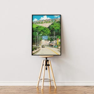 May include: A framed print of a road scene with a car driving towards a sign. The artwork features a road leading into a town, with lush green trees and a blue sky. The print is displayed on a wooden stool.