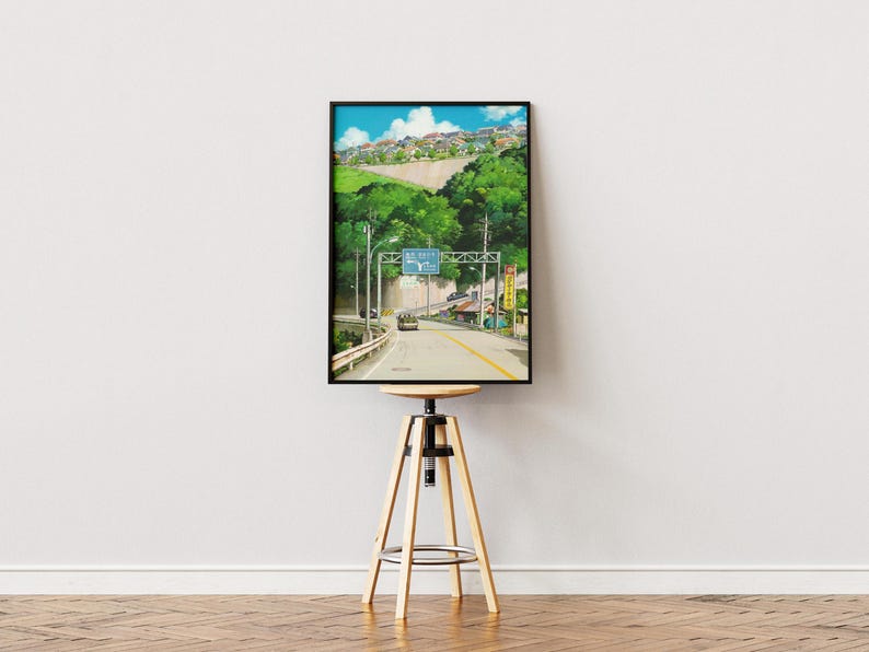 May include: A framed print of a road scene with a car driving towards a sign. The artwork features a road leading into a town, with lush green trees and a blue sky. The print is displayed on a wooden stool.