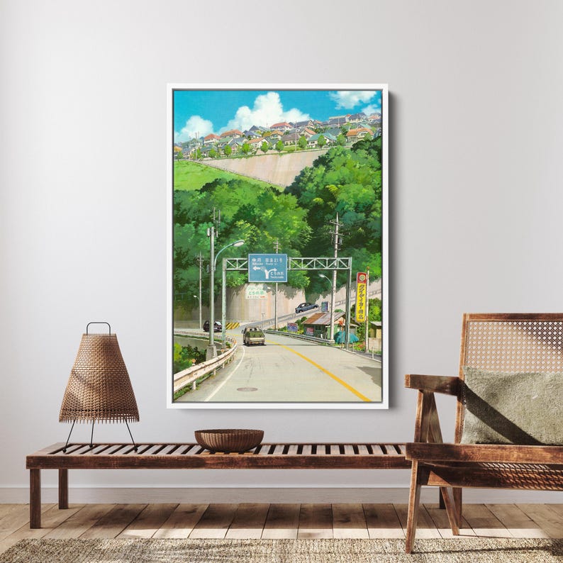 May include: A framed art print depicts a winding road through a lush, green landscape under a blue sky with fluffy clouds. A road sign and buildings are visible. The artwork is displayed on a white wall above a wooden bench.