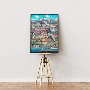 May include: A framed art print depicting a colorful cityscape with buildings, red roofs, and a harbor. The artwork is displayed on a wooden stool against a white wall. The print has a black frame.