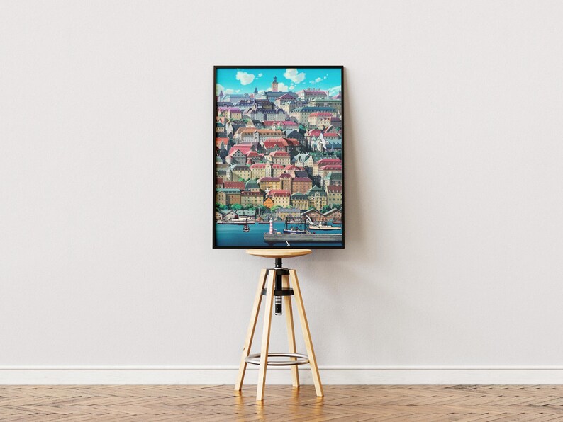 May include: A framed art print depicting a colorful cityscape with buildings, red roofs, and a harbor. The artwork is displayed on a wooden stool against a white wall. The print has a black frame.