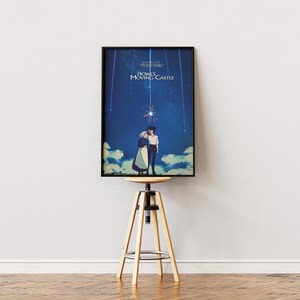 K&ouml;nnte beinhalten: Ein gerahmtes Poster mit dem Anime-Film "Das wandelnde Schloss". Das Kunstwerk zeigt zwei Figuren, die sich unter einem Sternenhimmel umarmen. Das Poster wird auf einem verstellbaren Hocker aus Holz und Metall vor einer wei&szlig;en Wand pr&auml;sentiert.