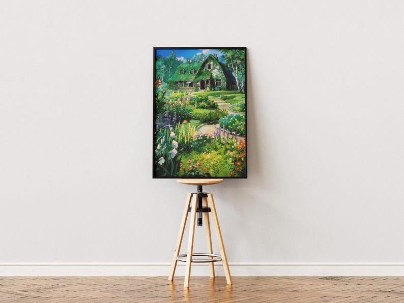 May include: A framed print of a picturesque garden scene. The artwork depicts a house with a green roof surrounded by lush greenery, colorful flowers, and a winding path. The print is displayed on a wooden stool.