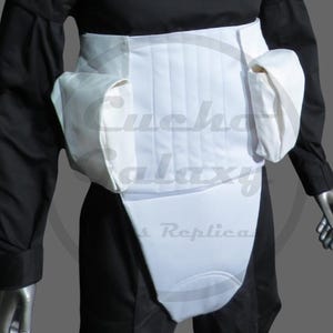May include: White costume belt with two pouches and a front panel, worn over a black outfit. The belt has vertical stitching and a curved lower section. The image shows a close-up of the belt, with the text "Cucho Galaxy Replicas" visible.
