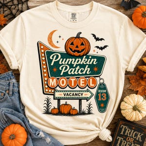 Puede incluir: Una camiseta color crema con un gráfico de un letrero de motel retro que dice Pumpkin Patch MOTEL y VACANCY, con una etiqueta de llave colgante marcada ROOM 13. El diseño incluye una calabaza de Halloween, una luna creciente, murciélagos.