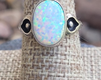 Könnte beinhalten: Silberring mit einem ovalen Opal-Edelstein mit schillernden Farben. Der Ring hat ein dekoratives Design auf jeder Seite des Opals. Der Ring wird auf einer strukturierten, neutralfarbenen Oberfläche präsentiert.