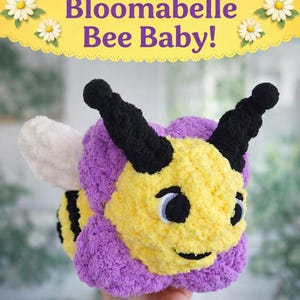 May include: A plush bee toy with a yellow face, purple body, and black antennae. The bee has a smiling expression and a white wing. The text "Bloomabelle Bee Baby!" is displayed above the toy.