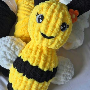 May include: A plush, handmade bee toy with yellow and black stripes. The bee has large, smiling eyes, a small orange flower, and white wings. The toy is made of soft, textured yarn, perfect for children.