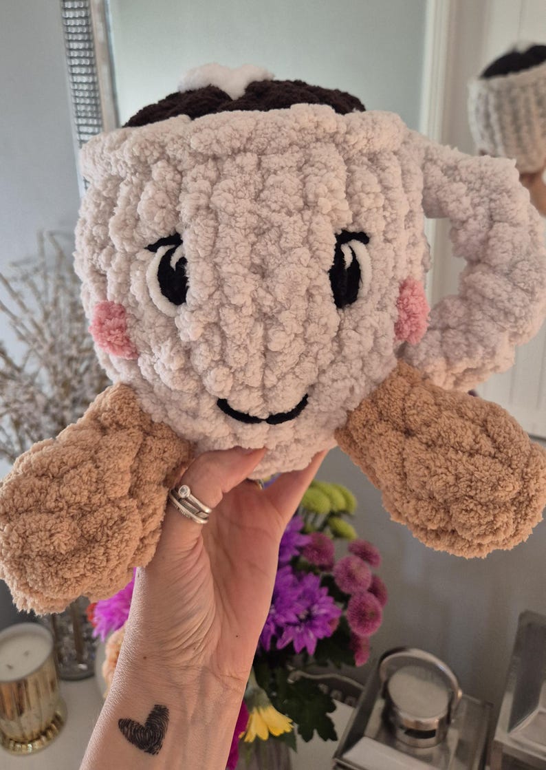 Pu&ograve; includere: Un peluche lavorato all'uncinetto a forma di tazza color crema, con una parte superiore marrone scuro, un accento di schiuma bianca e braccia color marrone chiaro. La tazza presenta occhi neri ricamati, un sorriso nero e guance rosa.
