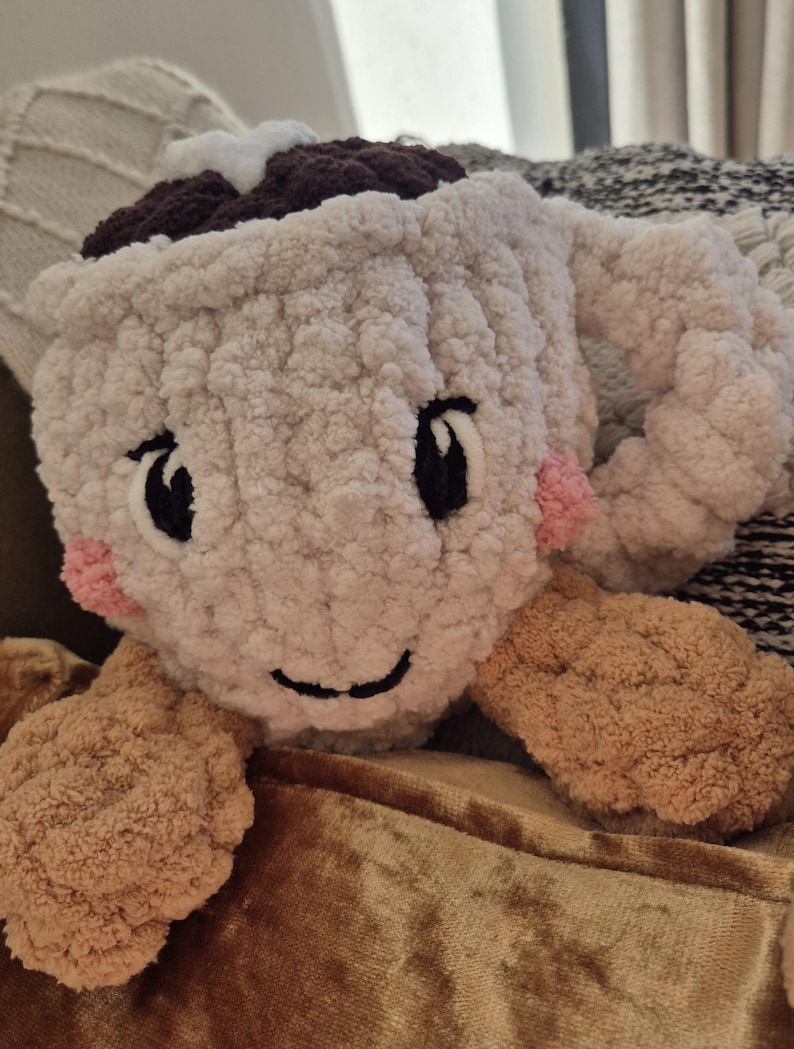 Pu&ograve; includere: Una tazza all'uncinetto in peluche color crema con una parte superiore marrone scuro, un accento a stella bianca e un manico. La tazza presenta occhi neri ricamati, un sorriso nero e piccole guance circolari rosa. Due appendici rotonde e testurizzate.