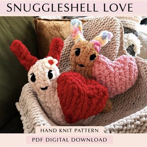 May include: Two plush snail-like creatures made of chunky yarn, one cream with red horns and a small flower, the other tan with multicolored horns. They are nestled in a cream-colored chunky knit basket with a red and a pink heart-shaped pillow.