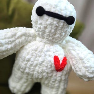 May include: A handmade white plush toy with a red heart-shaped detail on the chest. The toy has black eye-shaped accents and a black bar across the eyes. The plush toy is made of a soft, textured material.