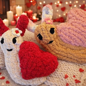 May include: Two plush snail-shaped toys with heart-shaped shells. One is cream-colored with a red heart, the other is tan with a pink heart. Both have black eyes and smiling faces. The background includes candles and a fireplace.