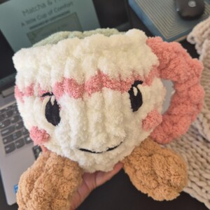 Pu&ograve; includere: Un peluche all'uncinetto a forma di tazza con corpo bianco, bordo a righe rosa e manico rosa. Presenta occhi ricamati neri, un sorriso nero e guance rosa. La base &egrave; color marrone chiaro con due piccoli piedi arrotondati sul fondo.