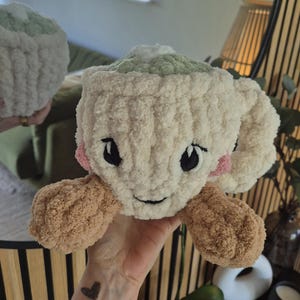 Pu&ograve; includere: Una tazza in crochet di peluche color crema con una parte superiore verde chiaro, due braccia in crochet marroni e guance rosa. La tazza presenta occhi neri ricamati e una piccola bocca nera, creando un volto amichevole. La texture &egrave; morbida.